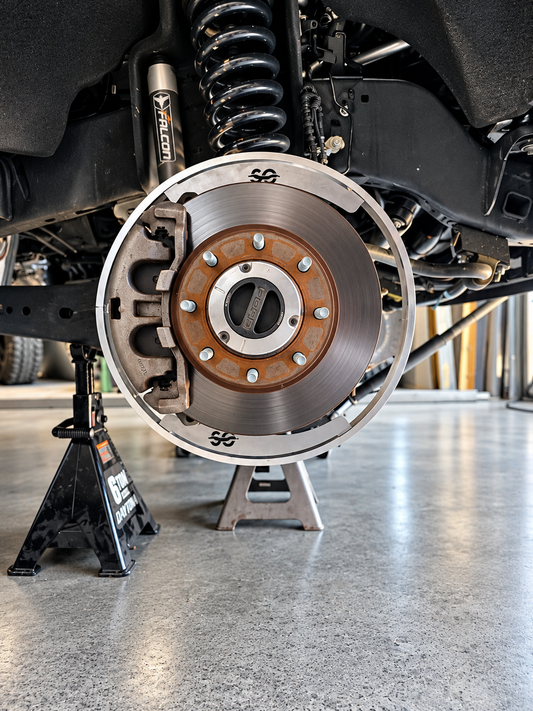 Shoreline Offroad 2N1 removable wheel light bracket ring fitted around a brake disc with the wheel removed, vehicle on jack stands in a shop